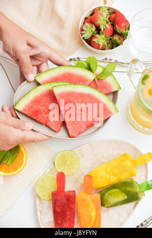 junger Mann, eine Platte mit einigen Scheiben Wassermelone auf einem Tisch platzieren wo gibt es einige verschiedene hausgemachte natürliche Eis am Stiel in einem Teller mit Eis, und ein Stockfoto