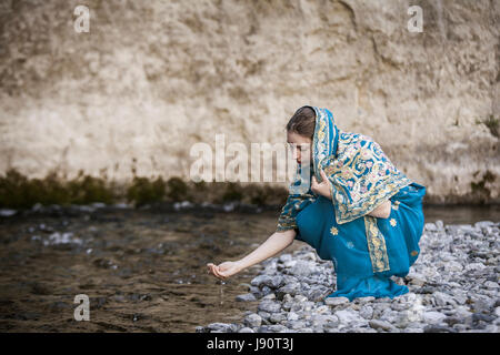 Das Mädchen in die indischen Sari sitzt an dem kleinen Fluss und trinkt aus Händen Stockfoto