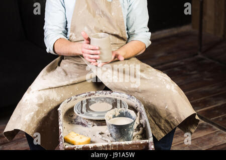 Keramik, Werkstatt, Keramik-Kunst-Konzept - close-up auf männlichen Händen mit ungebrannten Ton Cup, ein Mann untersucht ein frisches Produkt vor der weiteren Verarbeitung, Meister sitzt an der Töpferscheibe, Vorderansicht. Stockfoto