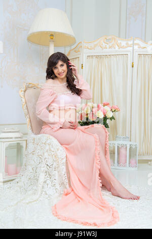 Schöne Mutterschaft. Schwanger in zarten Pfirsich Kleid sitzt auf Stuhl Stockfoto