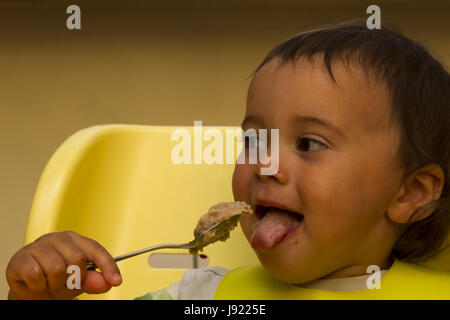 Happy Baby mit Löffel im Hochstuhl sitzt und isst Brei auf Platte. Geringe Schärfentiefe. Stockfoto