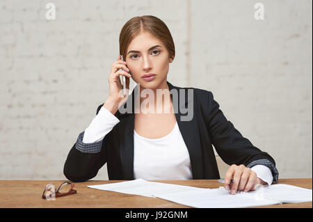 Schönen Business-Dame am Telefon zu sprechen. Sie prüft die Berichte und hält den Stift. Brille neben ihr. Stockfoto