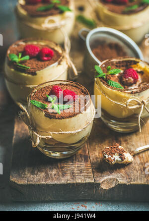 Hausgemachte italienische Dessert Tiramisu in einzelne Gläser mit Minze, frische Himbeeren und Kakaopulver auf rustikalem Holzbrett über graue c serviert Stockfoto