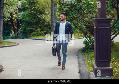 Stilvollen Mann mit einem Bart und Sonnenbrille geht in den Park und trägt eine Tasche. Stockfoto