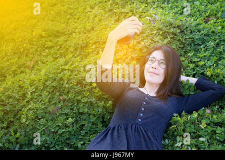 Asiatische Frau Lächeln Gesicht Selfie und sitzen auf dem Rasen. junge Asiatin Selfie auf glückliche Zeit. Konzept der entspannende Reise. Selektiven Bildschärfe auf fa Stockfoto