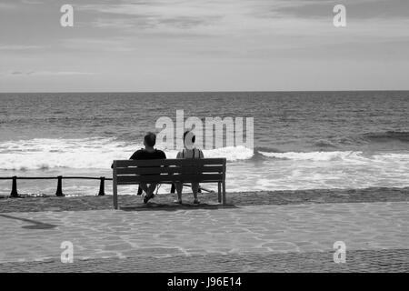 Urlaub paar sitzen und entspannen auf einer Holzbank, wie sie in den Atlantischen Ozean von der Promenade am Playa Las Americas in Teneriffa in th aussehen Stockfoto