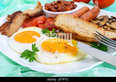 Traditionelles englisches Frühstück: Speck, Pilzen, Eiern, Tomaten, Würstchen, Bohnen, Toast auf einem weißen Teller auf einem hellen Hintergrund aus Holz.  England-Klasse Stockfoto