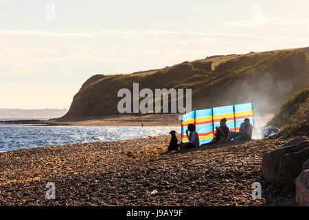 Hintergrundbeleuchtete Anzeigen einer Familie genießen Sie einen abendlichen Barbecue am Strand, in der vor einem bunt gestreiften Windschutz silhouetted, Ringstead Bay, Dorset, Großbritannien Stockfoto