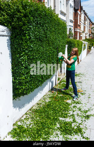 12-jährige Mädchen in einem grünen T-Shirt Schneiden eine buschige Hecke in einer Wohnstraße mit einer elektrischen Heckenschere Stockfoto