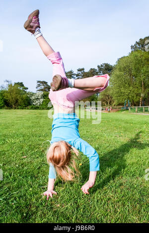 Sechs oder sieben Jahre alten Mädchen in einem Oben blau und rosa Jeans erfasst Mitte Sprung beim Handstand und Wagenräder auf dem Gras in einem Park Stockfoto