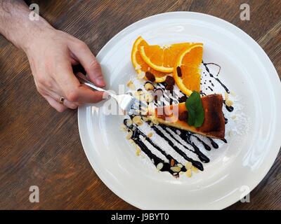 Mannes Hand mit einer Gabel mit Stück Käsekuchen mit Scheiben von orange und Schokoladensauce, Rosinen, Mandeln und Nüssen. Stockfoto