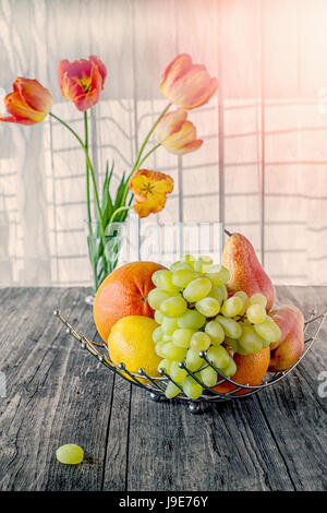 Frucht, Birne, Grapefruit, Zitrone, Pfirsich, Erdbeere in einem Korb auf dem Hintergrund der einen Strauß Tulpen an einem sonnigen Tag. Stockfoto