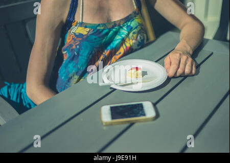 Eine junge Frau sitzt an einem Tisch draußen mit einem Scone und ein Handy Stockfoto