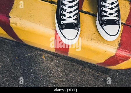 Schwarz / weiß neue Turnschuhe, Teenager Füße auf gelbe rote städtische Straße Grenze stehen. Nahaufnahme Foto mit selektiven Fokus Stockfoto