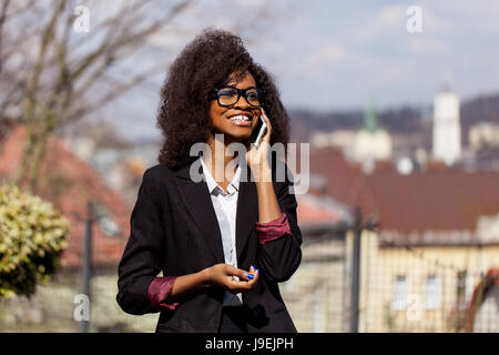 Lächelte schwarze Geschäftsfrau am Telefon zu sprechen. Frau mit dunklen Locken stehen auf dem großen Stadt Ansicht Hintergrund. Stockfoto