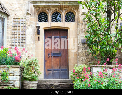 Alte dunkle braune Holztüren in Kalkstein-Hütte, umgeben von Blumen und Pflanzen. Stockfoto