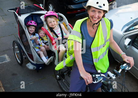 Frau Radfahrer auf Bike / Fahrrad mit + 3 Kinder; Co-Pilot Kindersitz mit Helm & Abschleppen Zyklus Chariot Anhänger mit zwei / 2 Kinder mit Helmen. VEREINIGTES KÖNIGREICH. Stockfoto