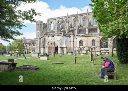 Eine Frau sitzen auf einer Bank auf dem Friedhof aus dem 7. Jahrhundert Malmesbury Abbey an einem sonnigen Tag in Wiltshire, Großbritannien Stockfoto