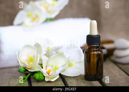 Eine Tropfflasche Bio Orchidee reines Öl auf eine Holzoberfläche. Weiße Handtücher, künstliche Orchidee Köpfe, rives Kieselsteine im Hintergrund. Stockfoto