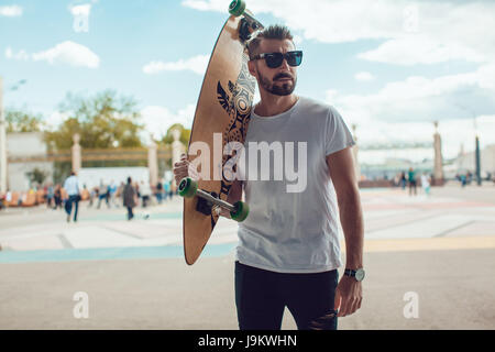 Stilvollen Mann mit Sonnenbrille und mit einem Bart steht auf der Straße mit einem langen Brett. -T-Shirt mock-up. Stockfoto