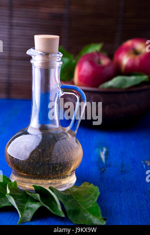 Apfelessig in Glasflasche auf blauem Hintergrund. Rote Äpfel in braunen Schüssel Stockfoto