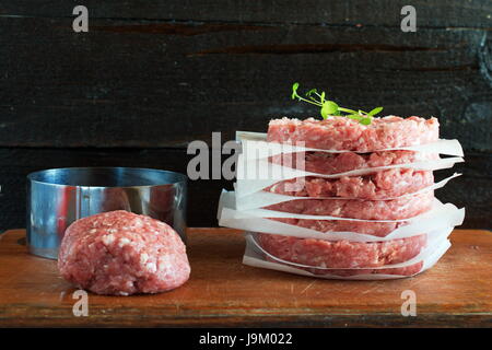 Stapel von hausgemachte Rindfleisch Burger auf ein Holzbrett mit irgendeiner Minsed Fleisch und Metall Form auf einem Hintergrund. Home gesunde Ernährung Stockfoto