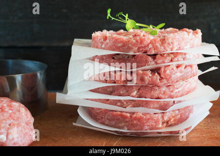 Stapel von hausgemachte Rindfleisch-Burger auf ein Holzbrett mit einigen gehackte Fleisch und Metall Form auf einem Hintergrund. Home gesunde Ernährung Stockfoto