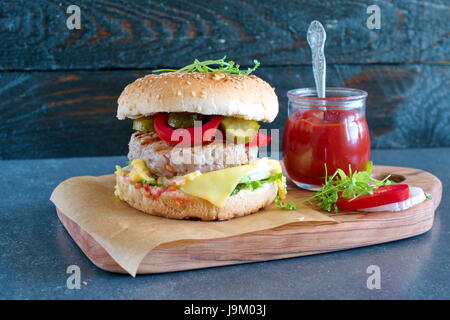 Hausgemachte Burger mit Brot, Käse, Tomaten, Salat, Tomaten-sauce, Gurken und Zwiebeln auf einem Holzbrett mit Glas mit Tomaten-sauce, Petersilie Stockfoto