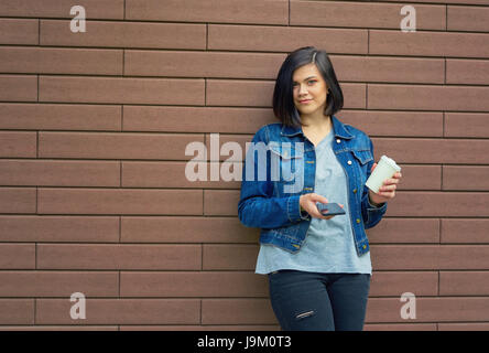 Schöne Brünette junge Frau mit Tunnel in den Ohren in eine blaue Jeans-Jacke mit einer Tasse Kaffee und Smartphone vor Mauer steht. Stockfoto