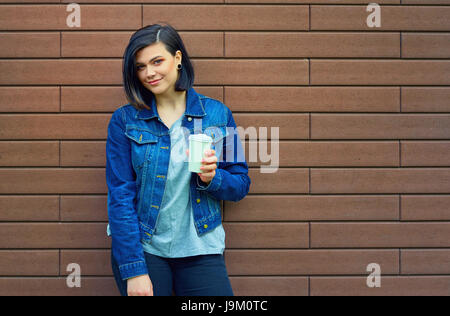 Hübsche Brünette junge Frau mit Tunnel in den Ohren in eine blaue Jeans-Jacke mit einer Tasse Kaffee vor Mauer steht. Stockfoto