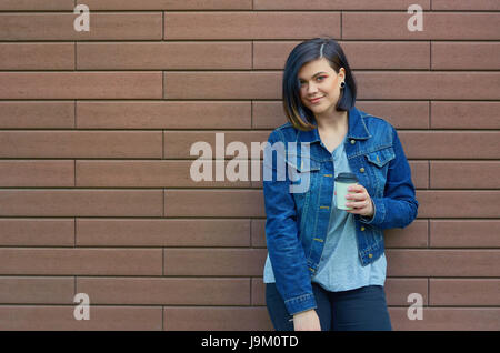 Schöne Brünette junge Frau mit Tunnel in den Ohren in eine blaue Jeans-Jacke mit einer Tasse Kaffee vor Mauer steht. Stockfoto