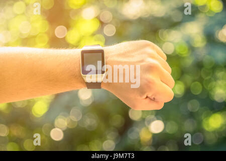 Nahaufnahme des männlichen Hand tragen Smartwatch mit leeren Bildschirm im Park. Grüne Blätter und Sonnenlicht Hintergrund unscharf. Gesunde Lebensweise und neue techn Stockfoto