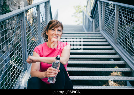 Glücklich und schön mittlere gealterte Frau sitzt auf metallischen Treppe Entspannung vor laufen im Freien halten Sie eine Flasche Wasser Stockfoto