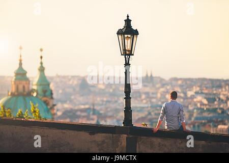 Junger Mann auf der Mauer sitzend und Skyline der Stadt bei Sonnenaufgang zu beobachten. Prag, Tschechische Republik Stockfoto