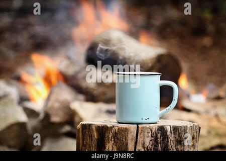 Blauer Emaille Tasse heißen dampfenden Kaffee auf eine alte Log ein Lagerfeuer im Freien sitzen. Extrem geringe Schärfentiefe mit selektiven Fokus auf Becher. Stockfoto