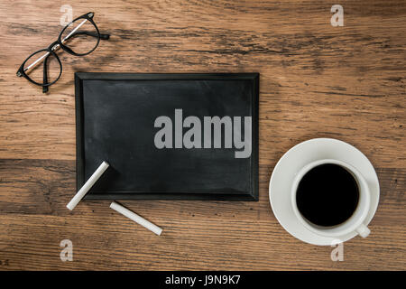 Tasse Kaffee auf einem hölzernen Schreibtisch-Hintergrund und eine leere Tafel mit zwei weiße Kreiden für Design Inspiration für Exemplar mit Stil Brille Stockfoto