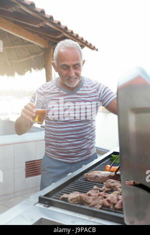 Kaukasischen Mann rund 60 Grillen von Fleisch und Gemüse auf ein Gas-Grill und genießen Sie ein Glas Bier Stockfoto