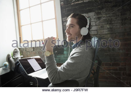 Lächelnd kreative Geschäftsmann mit Laptop Musikhören mit Kopfhörer und Füßen oben im Büro