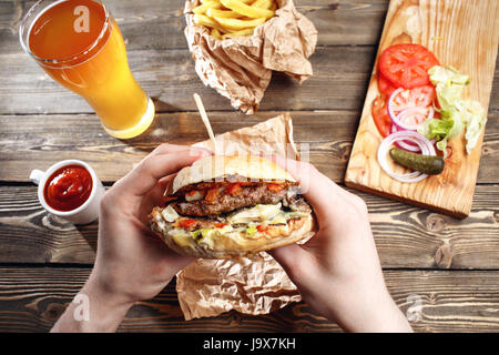 Hände halten frische leckere Burger mit Pommes frites, anzeigen Soße und Bier auf der hölzernen Tischplatte Stockfoto