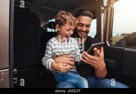 Schuss der glückliche Vater und Sohn sitzen auf der Rückseite des Autos und digital-Tablette zu betrachten. Junger Mann und kleiner Junge mit digitalen Tablet unterwegs Stockfoto