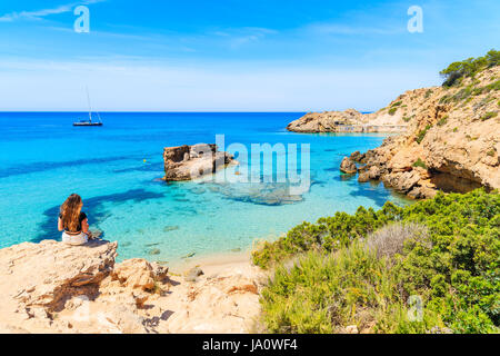 Unbekannte junge Frau sitzt auf einem Felsen und mit Blick auf schöne Cala Tarida Bucht, Insel Ibiza, Spanien Stockfoto
