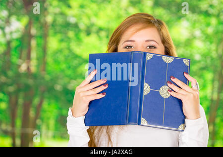 Kopfschuss junge Brünette deckt die Hälfte des Gesichts mit Blaubuch, Garten Hintergrund Stockfoto