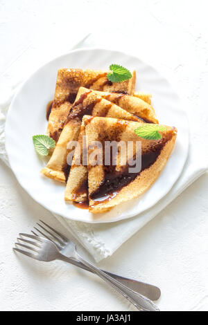 Crêpes mit Schokosauce und Minze auf weißen Teller auf weißem Hintergrund, Textfreiraum. Köstliche hausgemachte Pfannkuchen zum Frühstück. Stockfoto