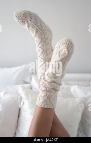 Weibliche Beinpaar Creme gemütliche Socken auf Bett liegend mit Füßen in der Luft mit weißen Kissen hinter Stockfoto