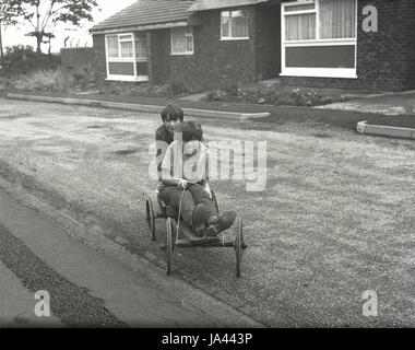 der 1970er Jahre, historische, zwei jungen, Reiten auf einem Haus gemacht aus Holz Go-kart mit Lenkung Seil aber keine Bremsen draußen spielen auf einem unmarkierten Straße. Stockfoto