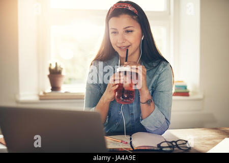 Junge lächelnde Geschäftsfrau mit leckeren Drink sitzen und mit Blick auf Laptop Kopfhörer. Stockfoto