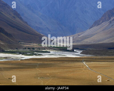 Hochtal, im Vordergrund eine riesige gelbe Wüste mit einem Faden von der Straße, im Hintergrund ein Fluss unter steilen Ufern, den Himalaya. Stockfoto