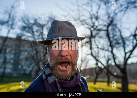 Ein bärtiger Mann mit Hut schreit vor Überraschung. Er ist draußen im Park spazieren. Stockfoto