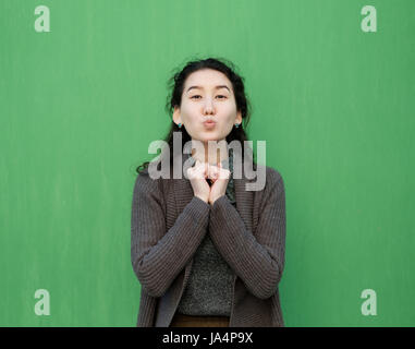 Schöne Asiatin sendet einen Luft-Kuss. Sie steht in einen warmen Pullover in der Nähe der grünen Wand Stockfoto