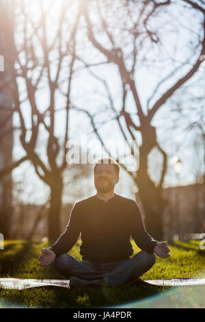 Reifer Mann sitzen auf Yoga-Matte in komfortablen Asana lächelnd und entspannen im Park. Meditation in Stadt Lebensbedingungen Stockfoto
